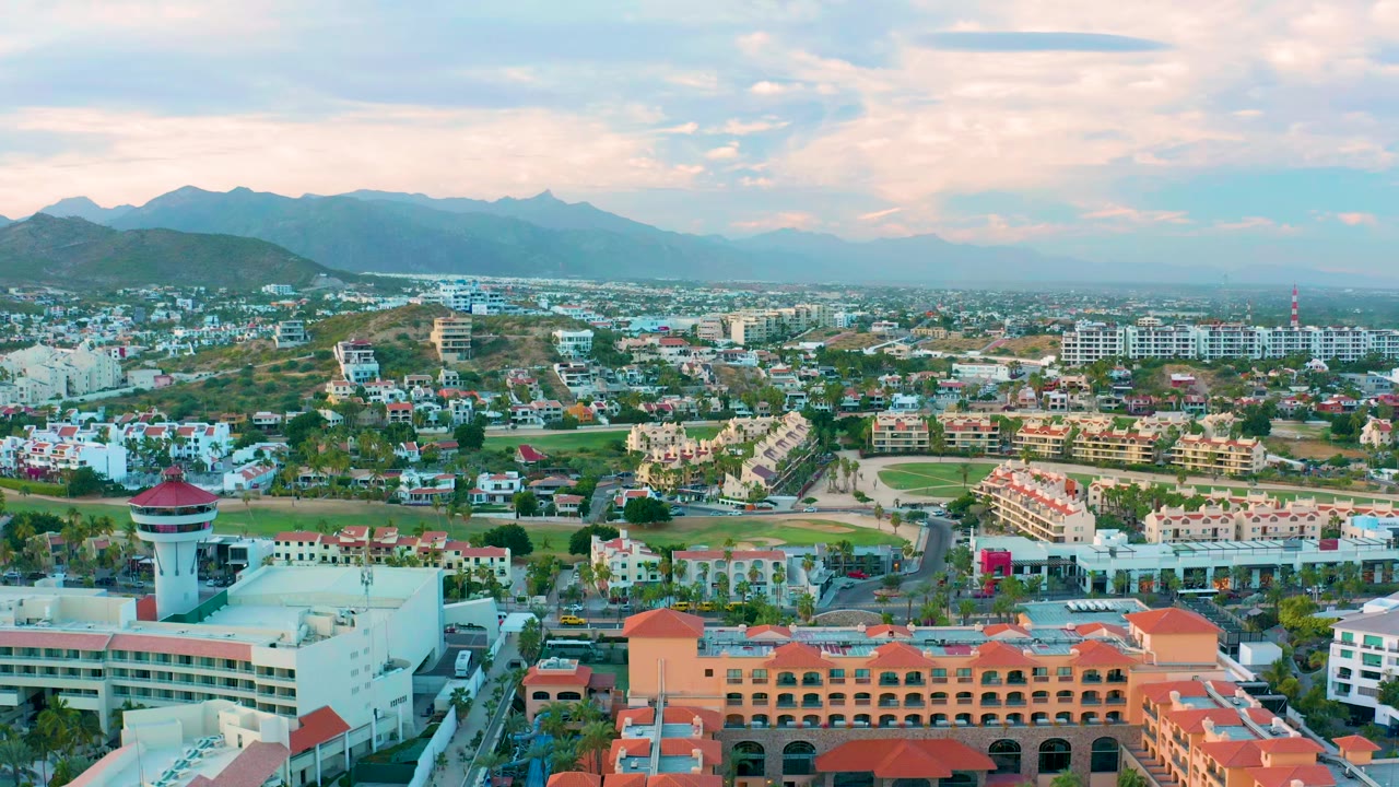 Wide aerial view over San Jose Del Cabo with red-roofed buildings in the foreground, low-rise neighborhoods across the middle distance, hazy mountains on the horizon, and no visible people.
