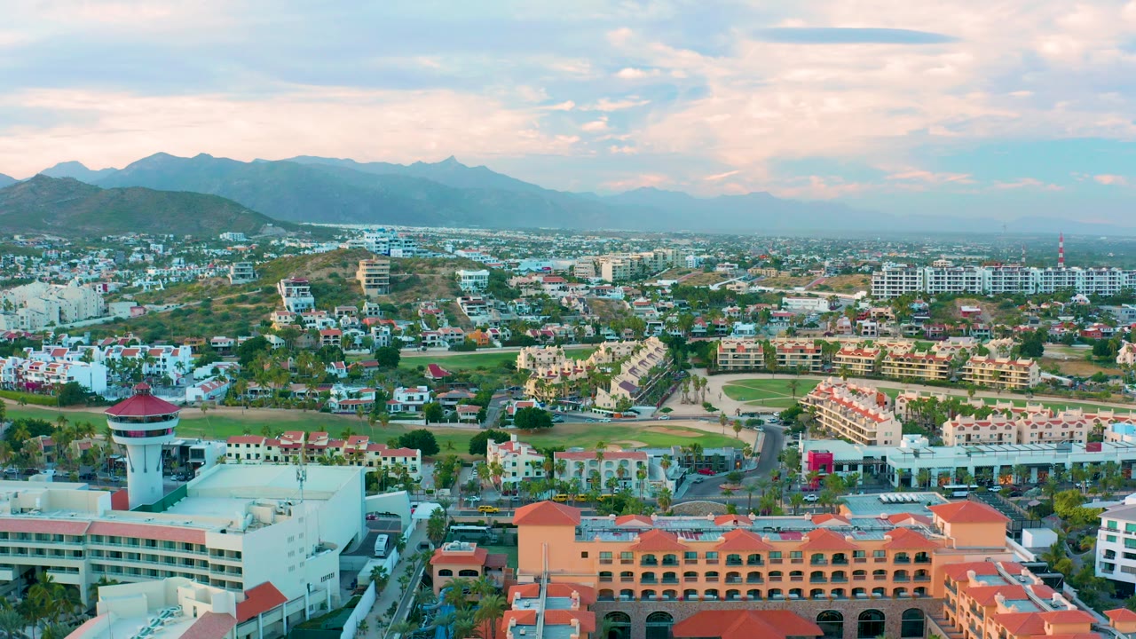 Aerial city view in San Jose Del Cabo with terracotta rooftops along the lower edge, compact residential and commercial buildings beyond, a hazy mountain range, pastel clouds, and no visible people.