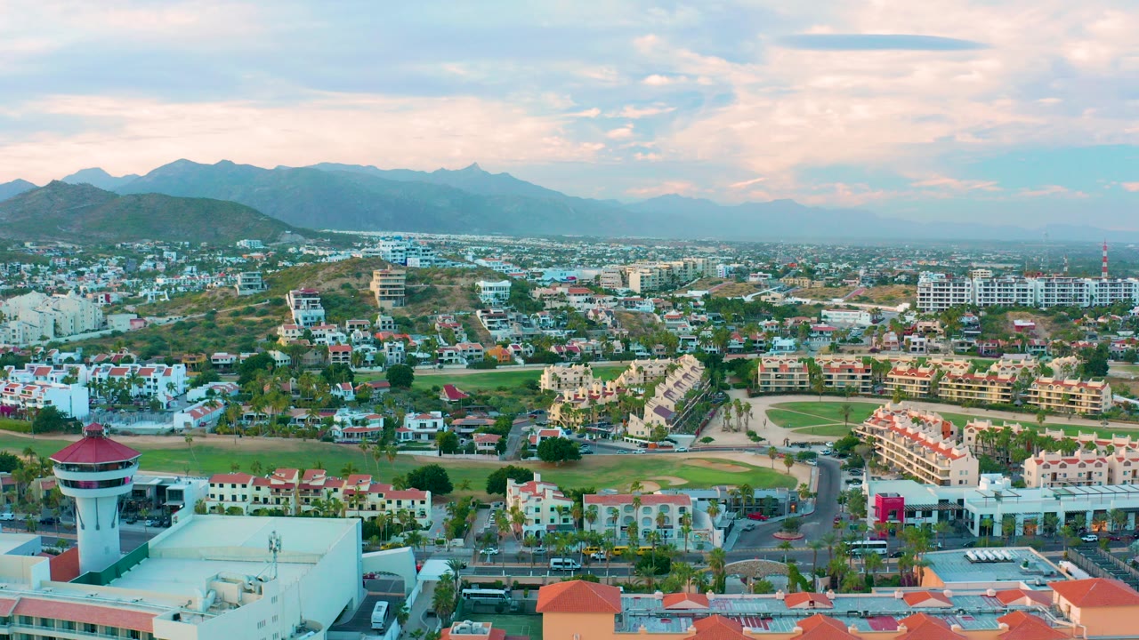 A wide aerial view over San Jose del Cabo shows rooftops, green field areas, roads, and a hazy mountain horizon under soft partly cloudy light, with no visible people.