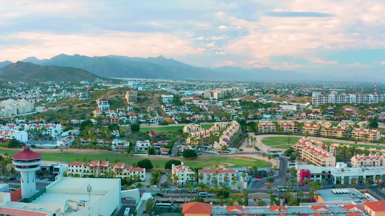 A high aerial frame over San Jose del Cabo shows dense low-rise buildings, a green field band, road lines, and layered mountains in the background, with no visible people.