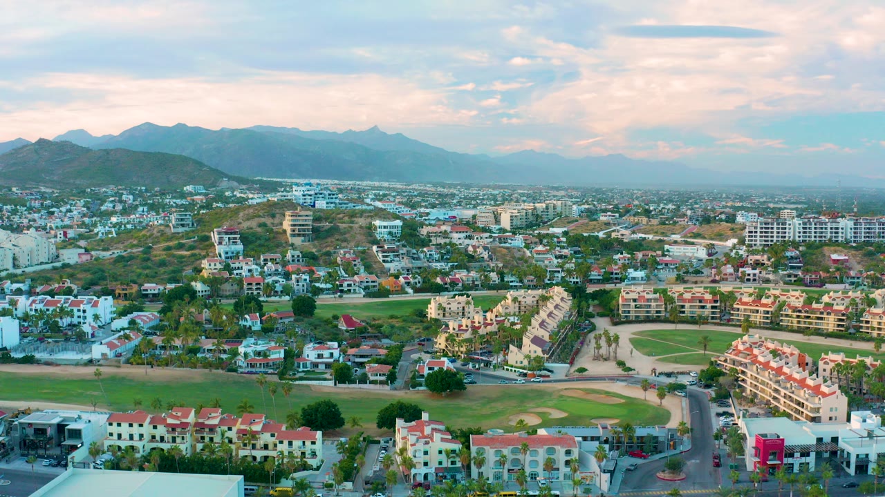 Opening aerial view over San Jose del Cabo with red-roofed residences, green open space, a road in the foreground, hazy mountains beyond, and no visible people.