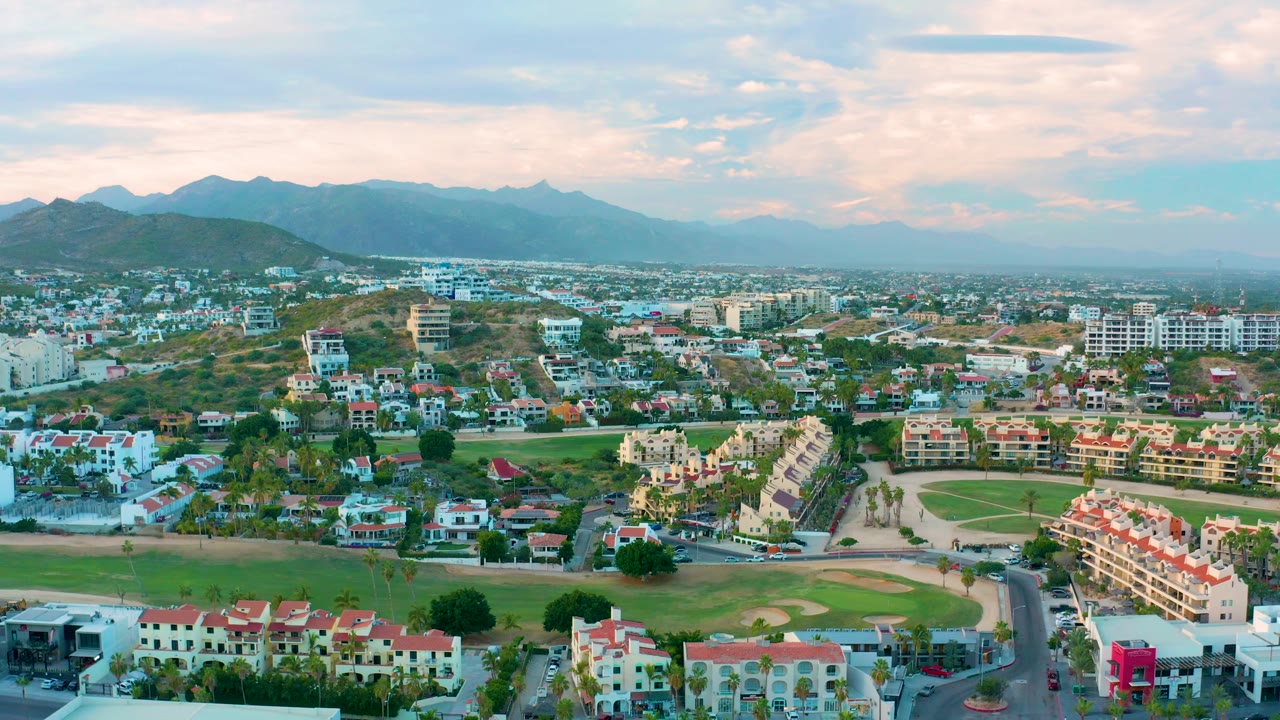 Aerial view across San Jose del Cabo residences, red-tile rooftops, open lawns, scattered trees, distant urban blocks, mountain ridges, and no visible people.