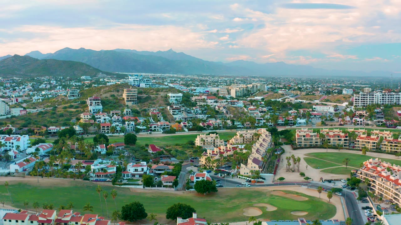 Aerial view over San Jose Del Cabo homes and a golf course with dry hills and blue mountains beyond, with no visible people.