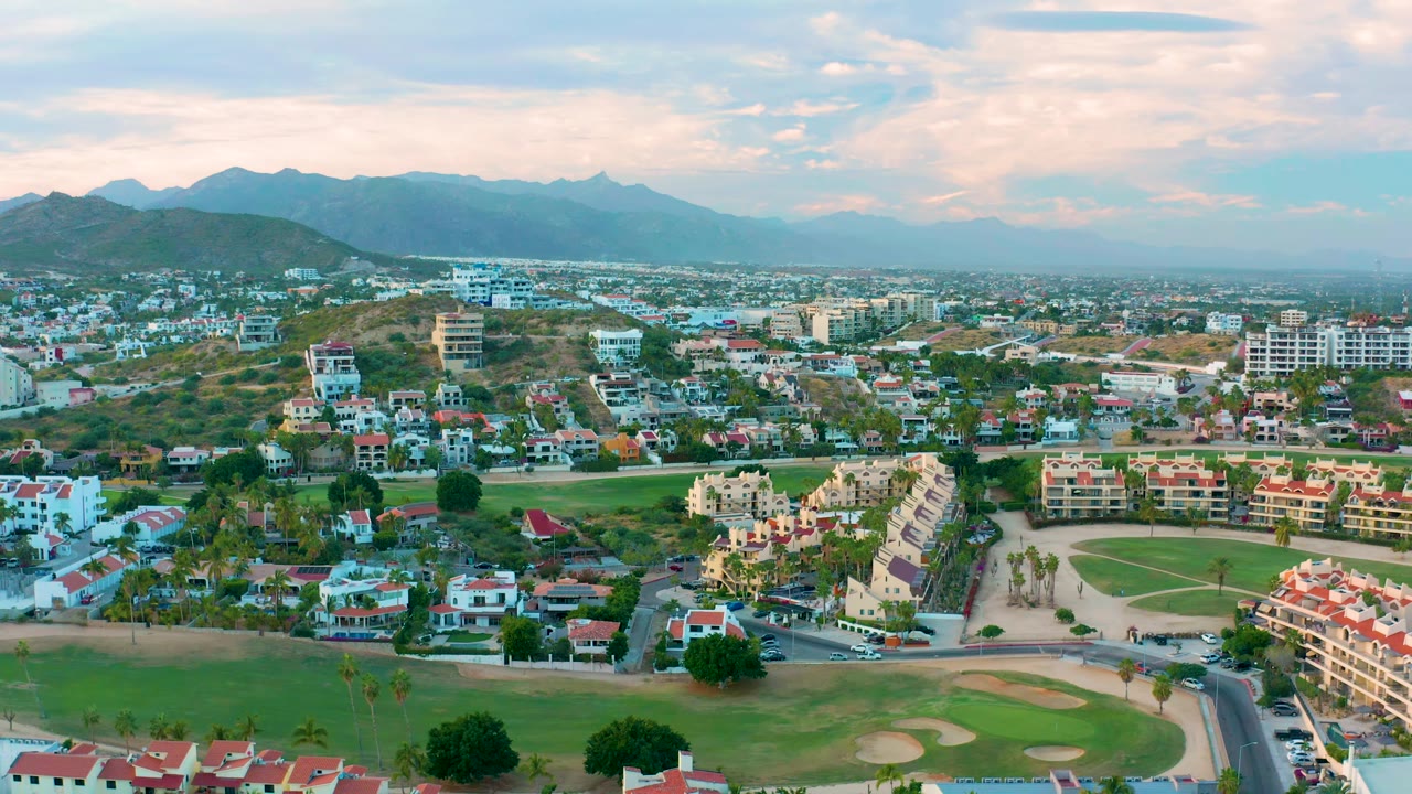 San Jose Del Cabo neighborhood seen from above with red-roof homes, a green fairway, road grid, and hazy mountains under warm clouds, with no visible people.