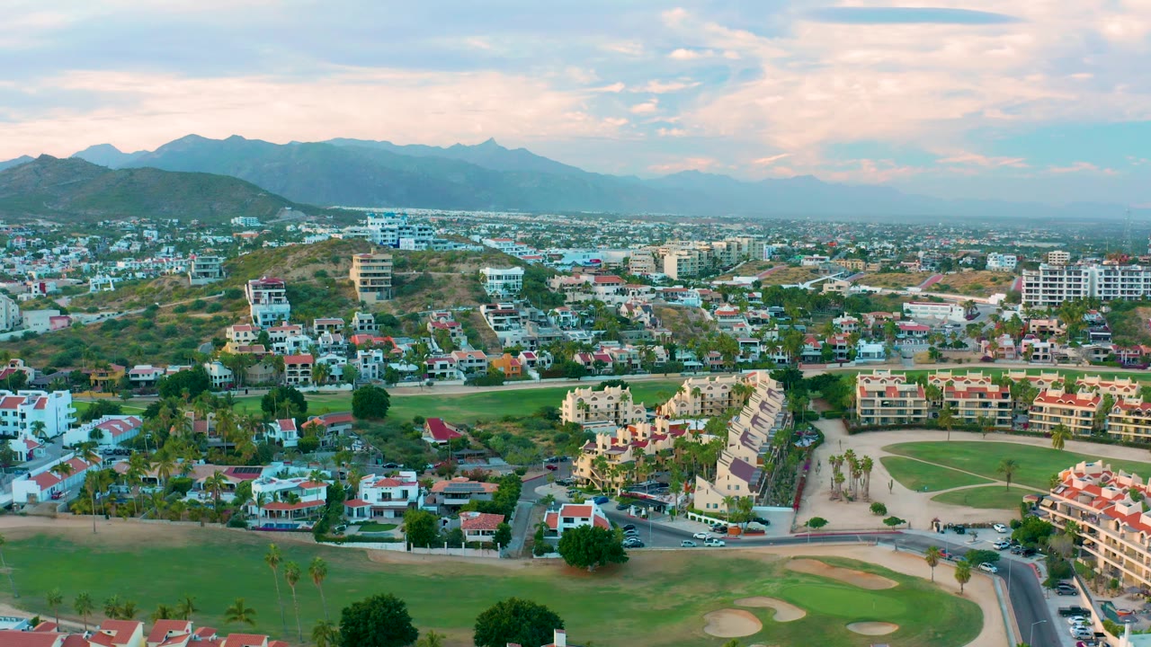 Wide San Jose Del Cabo aerial showing low-rise residences beside golf-course grass, open dry slopes, and layered mountain ridges, with no visible people.