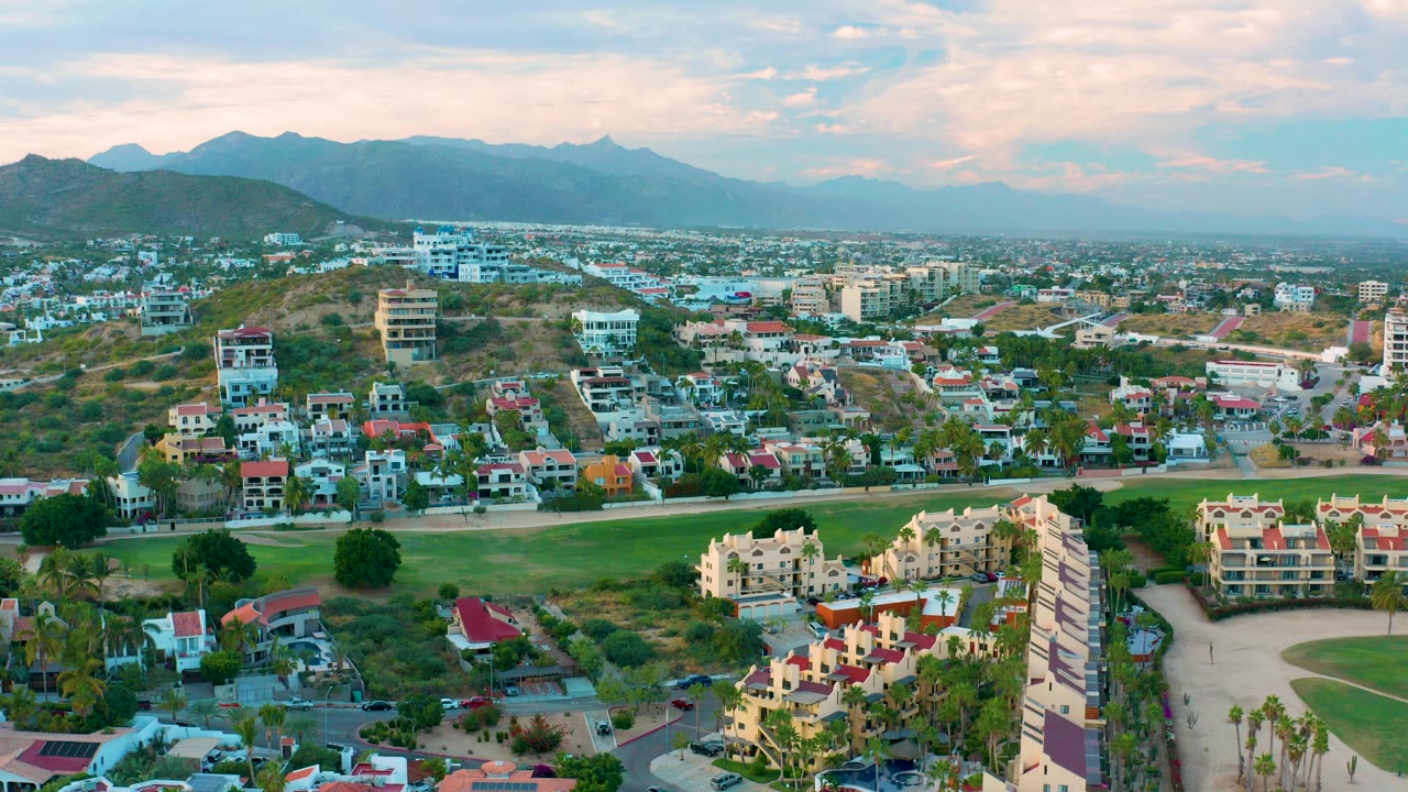 Opening aerial frame over San Jose Del Cabo residential hills, with a green field and road in the foreground, houses in the midground, mountains on the horizon, and no visible people.