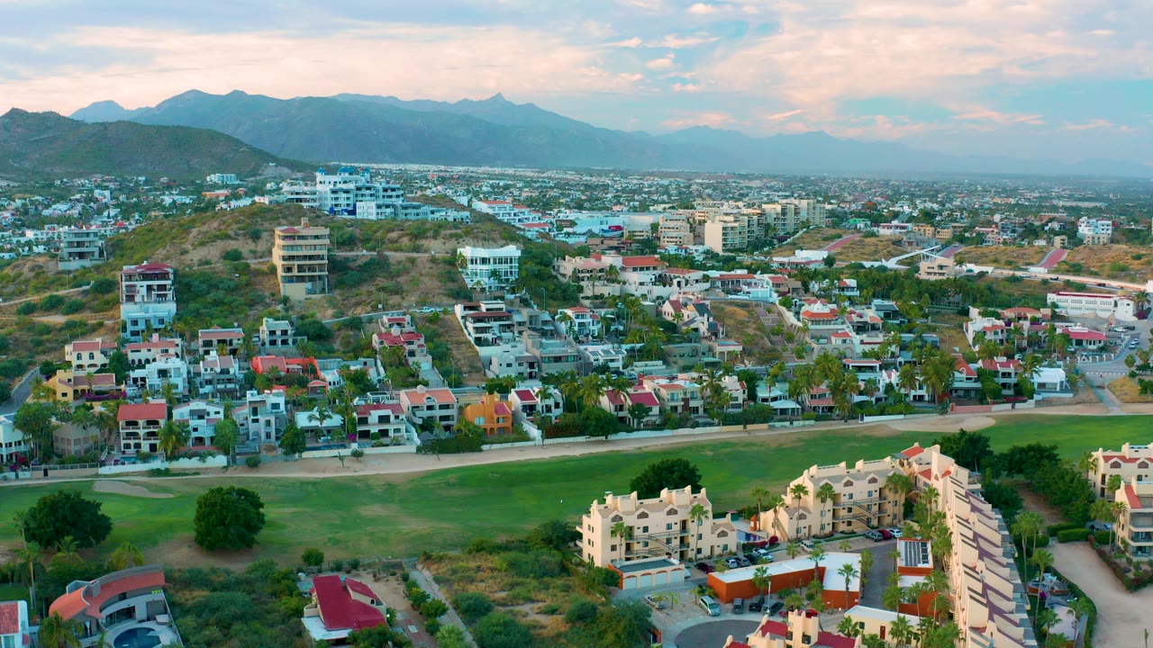 Aerial view over San Jose del Cabo with a green field at the bottom, houses and apartment buildings across the middle, distant mountains, pastel clouds, and no visible people.