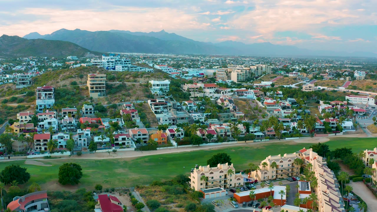 Aerial view in San Jose del Cabo with a road crossing in front of the homes, a green field in the foreground, scattered hillside buildings, mountain skyline, and no visible people.