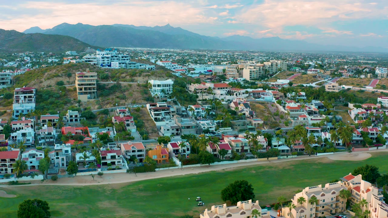 San Jose Del Cabo aerial frame looking over a green foreground, residential houses, hillside buildings, and distant mountains under warm clouds, with no visible people.