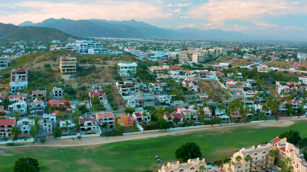 San Jose Del Cabo drone view with a broad green area at the bottom of frame, rows of houses and palms in the midground, hillside structures, and hazy mountains beyond, with no visible people.