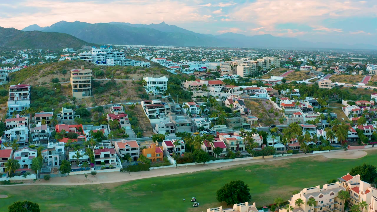 San Jose Del Cabo aerial frame showing red-roofed and light-colored homes, a curved road by green space, hillside lots, and a soft mountain horizon, with no visible people.