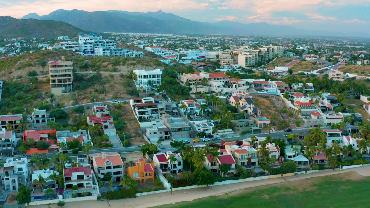 Aerial frame over San Jose Del Cabo showing beach sand at the bottom, colorful residential homes, hillside roads, dry terrain, distant mountains, pink clouds, and no visible people.
