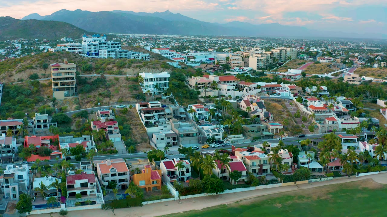 Aerial view in San Jose Del Cabo with the beach edge low in frame, a line of homes and palms beside the road, hillside buildings, town blocks beyond, distant mountains, and no visible people.