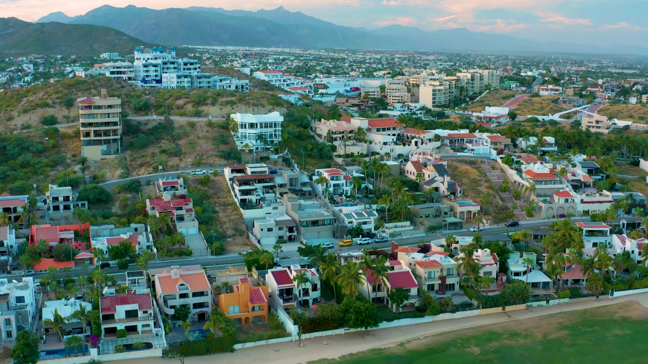 San Jose Del Cabo aerial frame showing a broader sweep of homes, a curving road with small cars, dry hills, dense town blocks, blue mountains, soft pink sky, and no visible people.