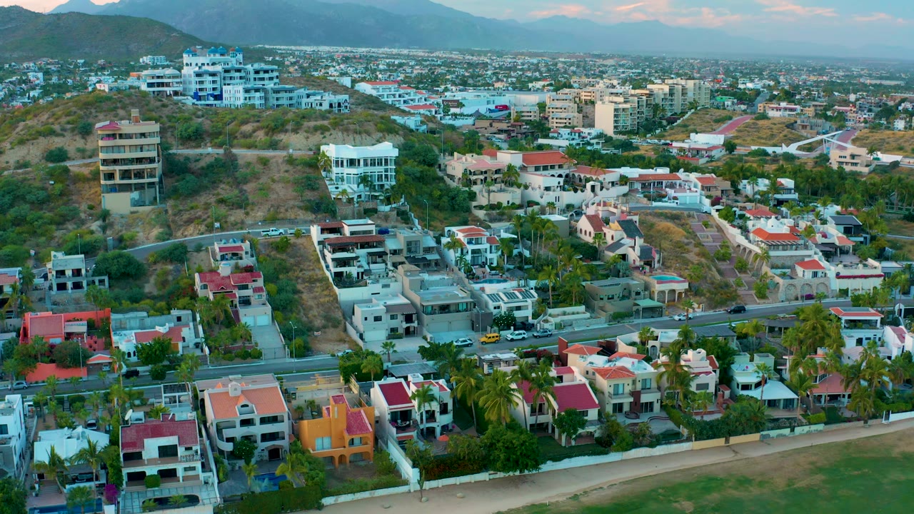 Aerial view over beachside residences, road traffic, dry hills, and distant city blocks.