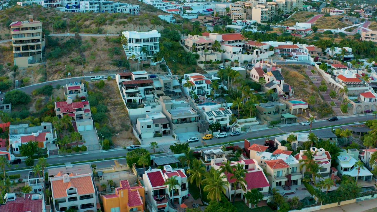 Aerial frame over San Jose Del Cabo homes with a paved road crossing the lower half, dry hillside behind the houses, red roofs, palms, parked vehicles, and no visible people.