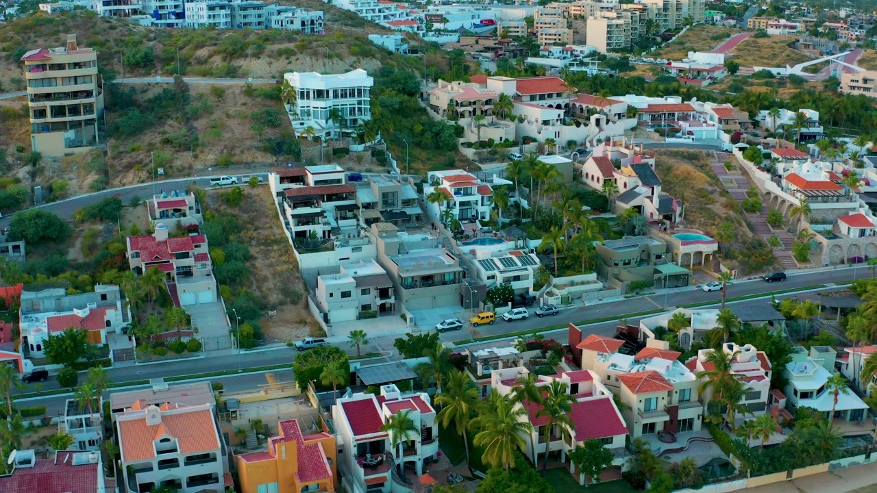High aerial view in San Jose Del Cabo showing flat-roof and red-roof houses, a road with several vehicles, palms, hillside lots, and no visible people.