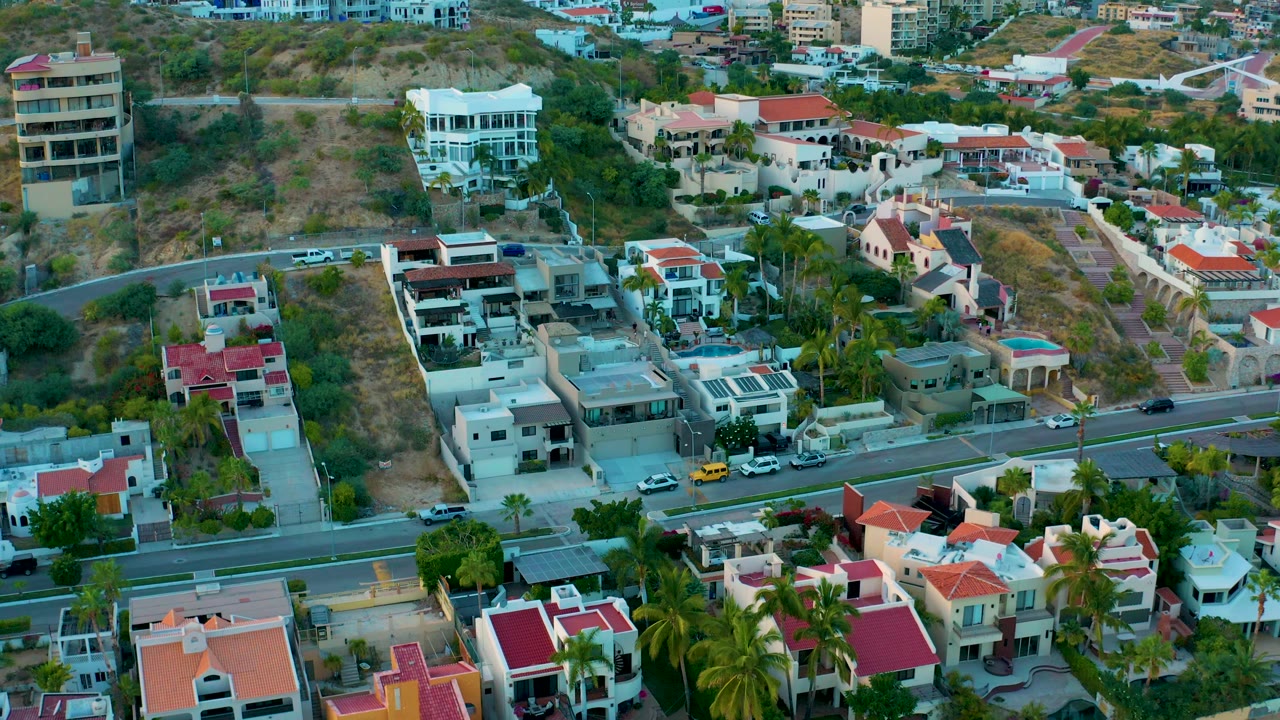 Aerial frame of a San Jose Del Cabo residential area with clustered homes, red tile roofs, a curving road, dry slopes, palms, parked cars, and no visible people.
