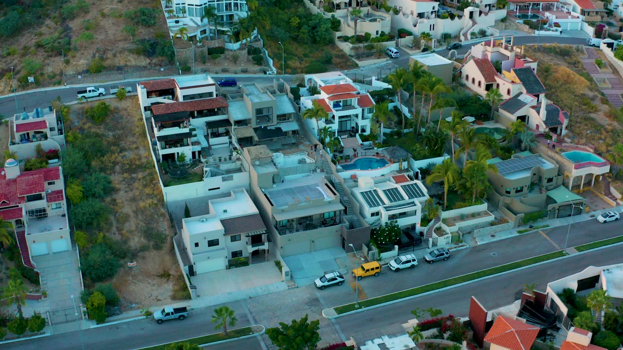 Drone view over San Jose del Cabo homes, parked cars, palm trees, a pool, and dry hillside terrain, with no visible people.