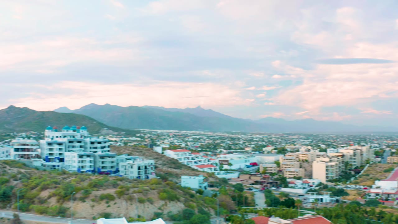 Elevated view of San Jose Del Cabo hillside buildings, a winding road, distant city blocks, mountain ridges, and soft pink-blue clouds, with no visible people.