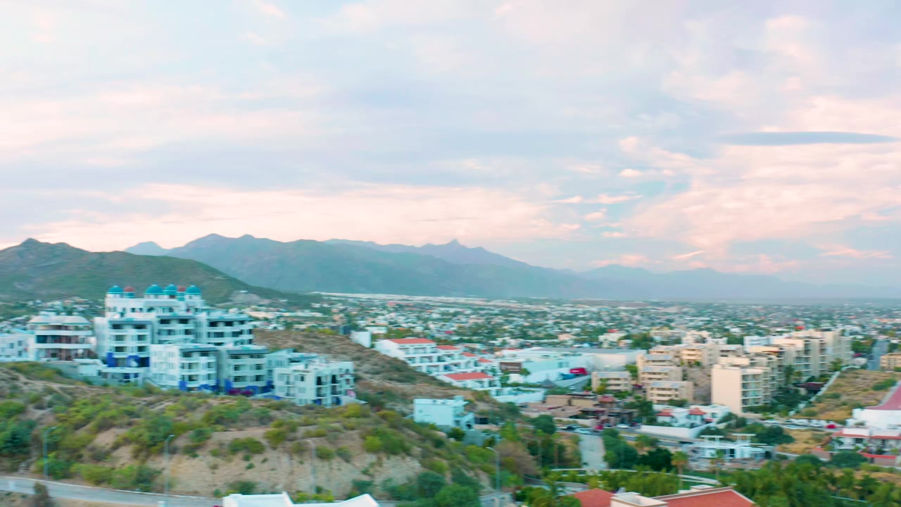 San Jose Del Cabo hillside development shifts slightly across frame, showing white multi-story buildings, green brush, valley buildings, and hazy mountain silhouettes with no visible people.