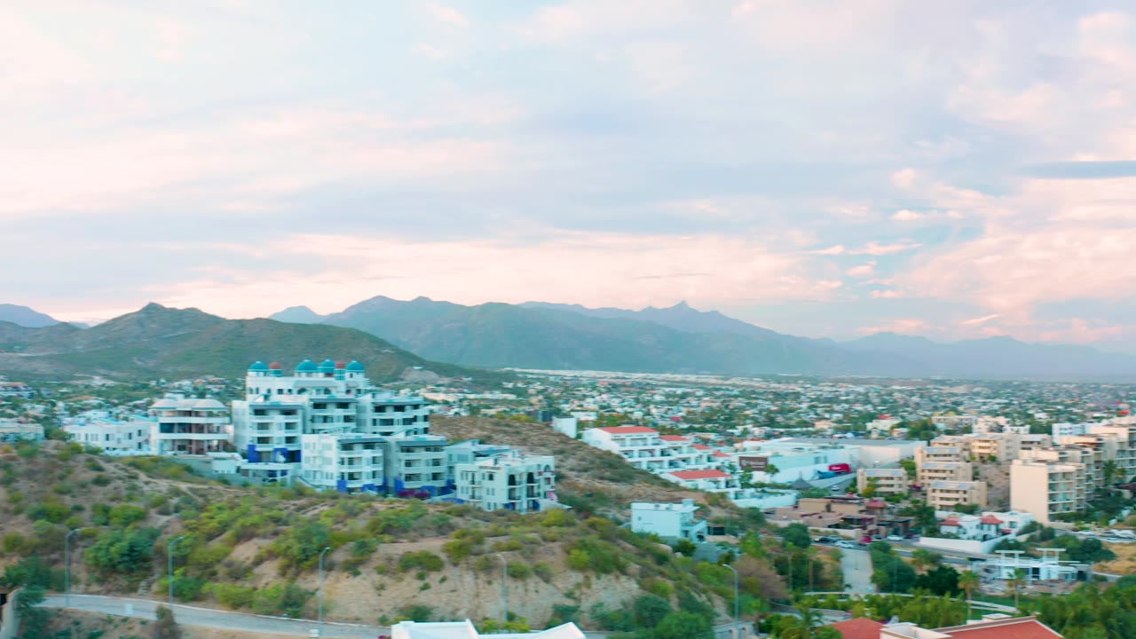 Elevated San Jose Del Cabo view with the white hillside buildings near center, low commercial and residential blocks across the midground, and layered mountains below pale evening clouds, with no visible people.