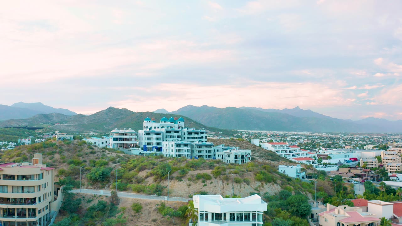 Wide aerial view of San Jose del Cabo hillside homes, dry green brush, a city below, distant mountains, pastel clouds, and no visible people.
