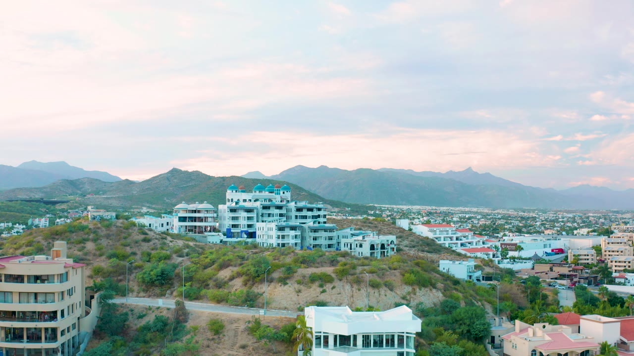 San Jose del Cabo aerial frame with a large white hilltop residence near center, tan buildings at the edges, city blocks beyond, mountains, and no visible people.