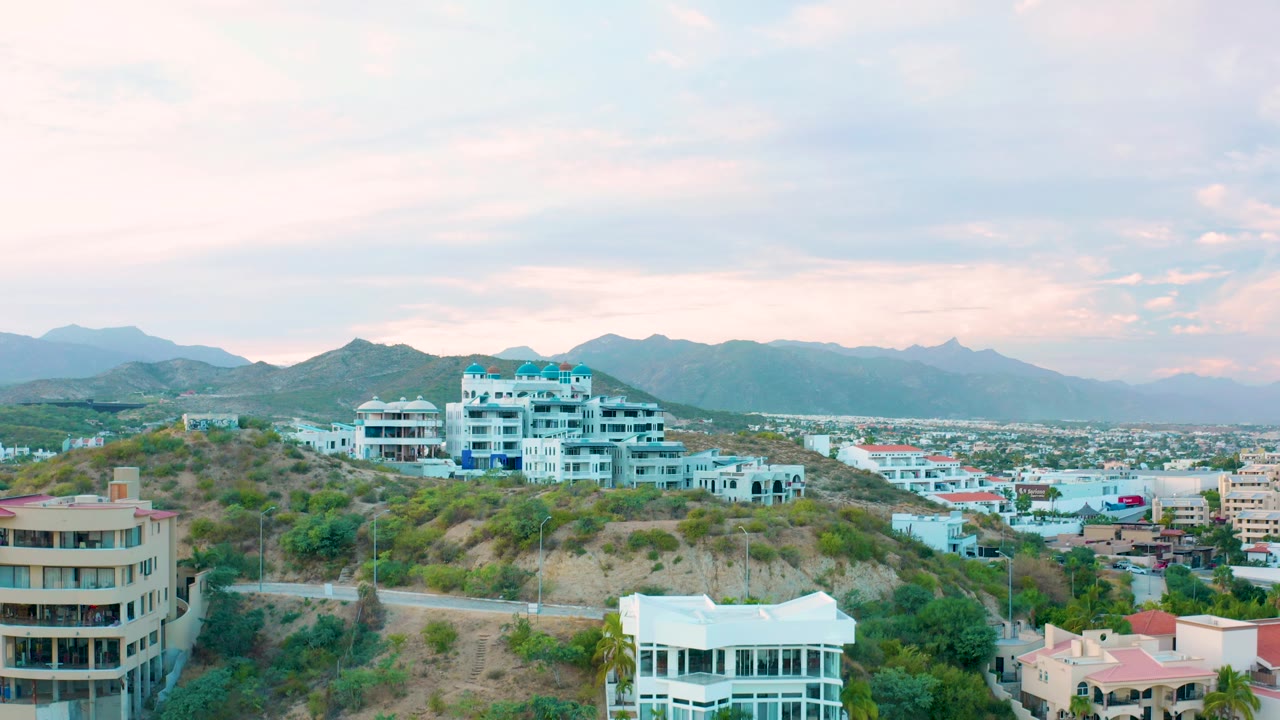 Aerial view in San Jose del Cabo showing white residential buildings on a brushy hill, a white foreground house, city development to the right, mountains, and no visible people.