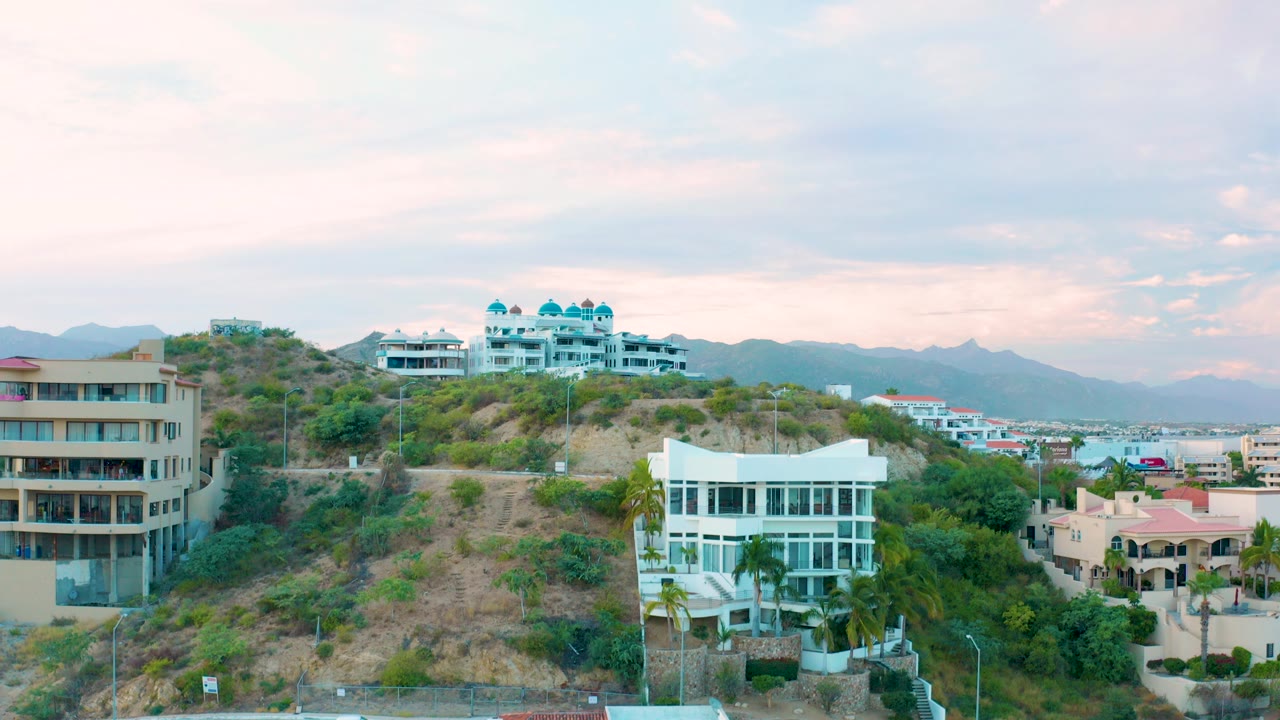 Elevated view of hillside residential buildings in San Jose Del Cabo with a white house, tan apartment block, distant mountains, soft clouds, and no visible people.