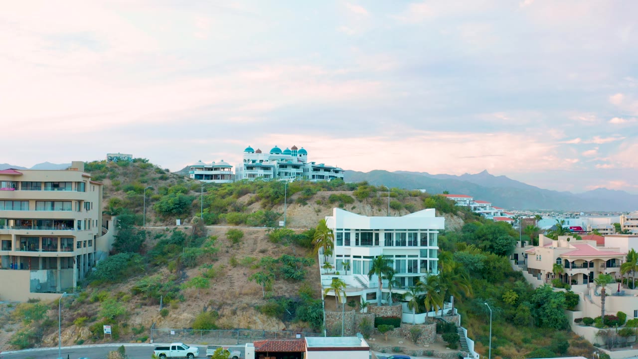 Residential hillside in San Jose Del Cabo with a white truck entering the lower edge, white and tan buildings on the slope, background mountains, and no visible people.