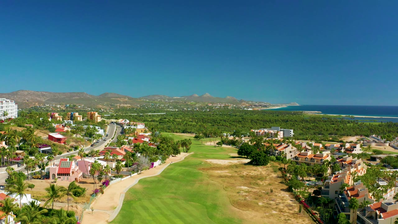 Wide aerial frame over San Jose del Cabo showing a green fairway, red-roof residential blocks, palm trees, distant mountains, and blue ocean at right, with no visible people.
