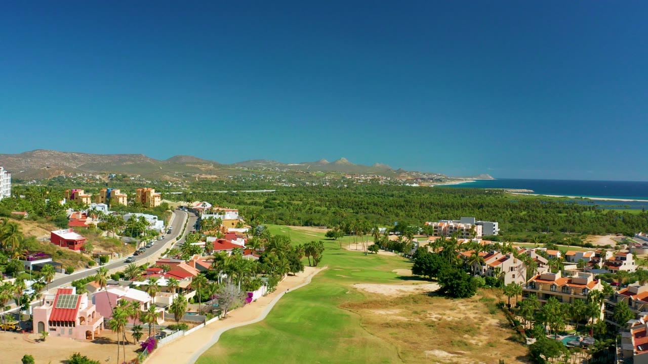 Aerial view in San Jose Del Cabo showing a curving road beside homes and golf greens, with mountains and ocean under a clear sky and no visible people.