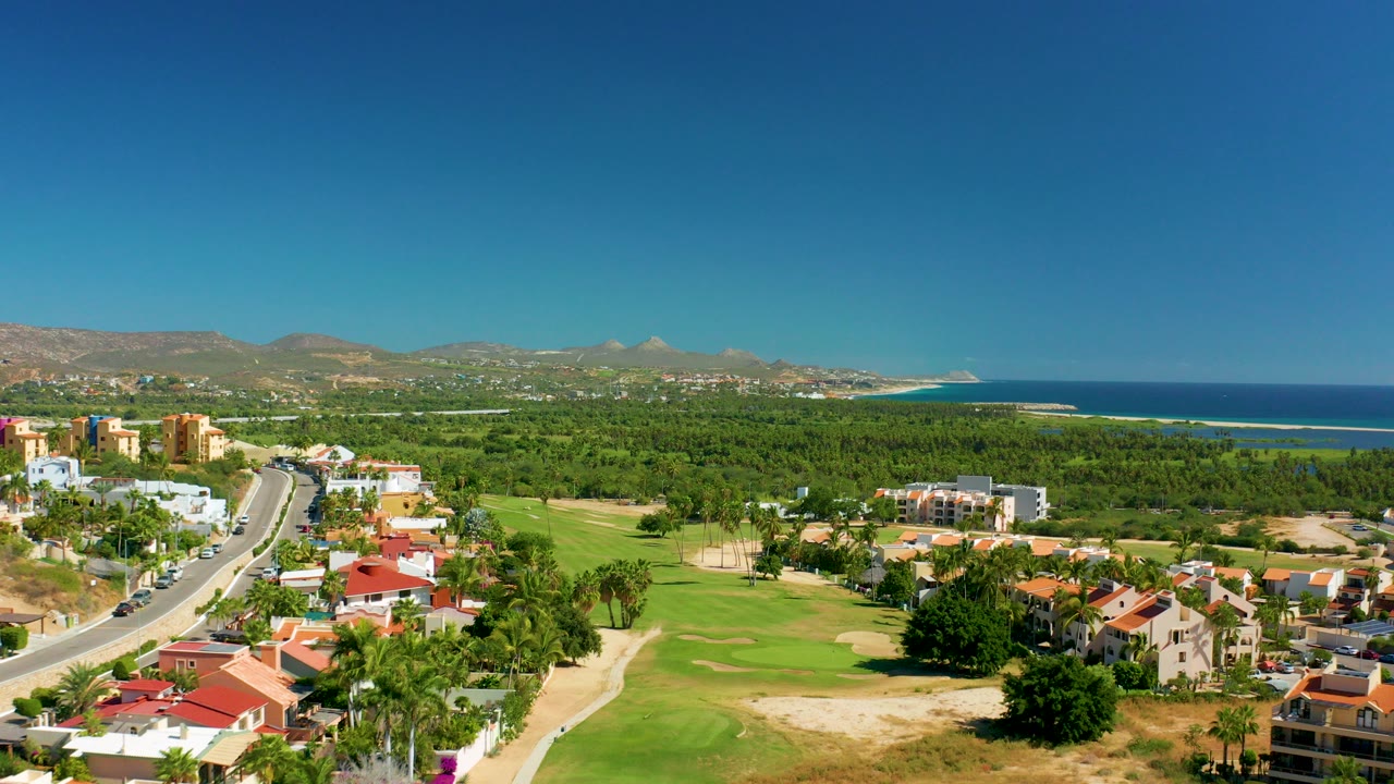 Aerial view of a curved road beside homes, golf greens, palm trees, mountains, and ocean.