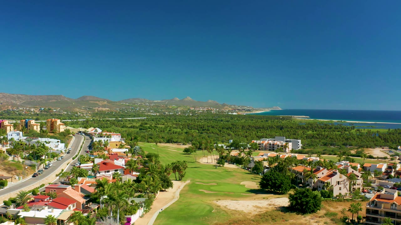 Second aerial moment in San Jose Del Cabo showing the road running between homes and golf greens, with the ocean and mountains across the background and no visible people.