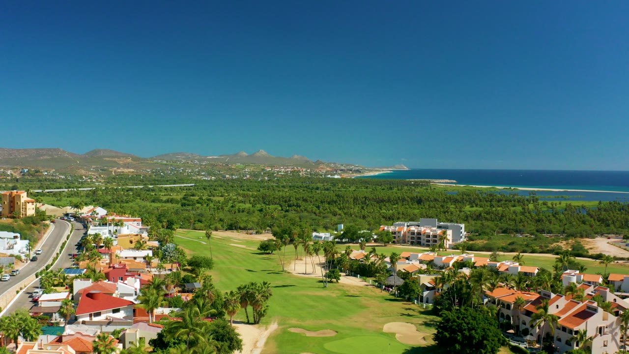 Opening aerial frame over San Jose Del Cabo showing a road at left, red-roofed homes, golf fairways, palm trees, ocean, distant mountains, and no visible people.
