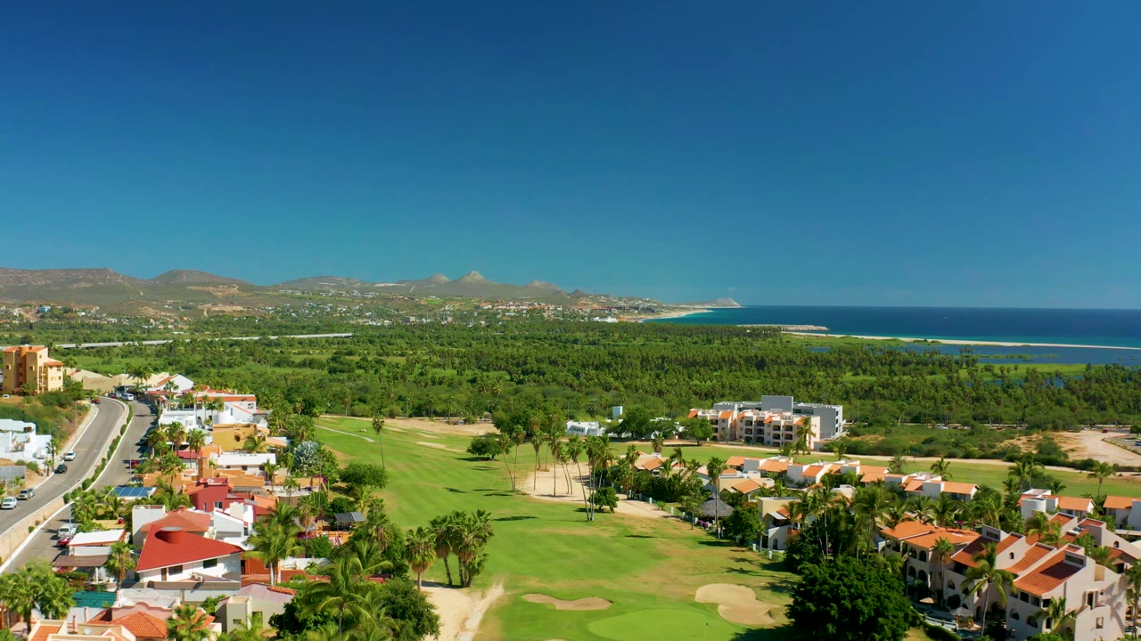 Aerial view in San Jose Del Cabo with the road running up the left side, green fairways in the foreground, homes below, blue ocean beyond the tree line, and no visible people.