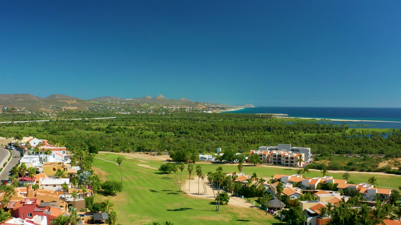 Opening aerial frame over San Jose del Cabo with green fairways and red-roofed homes in front of coastal trees, blue ocean, distant mountains, clear sky, and no visible people.