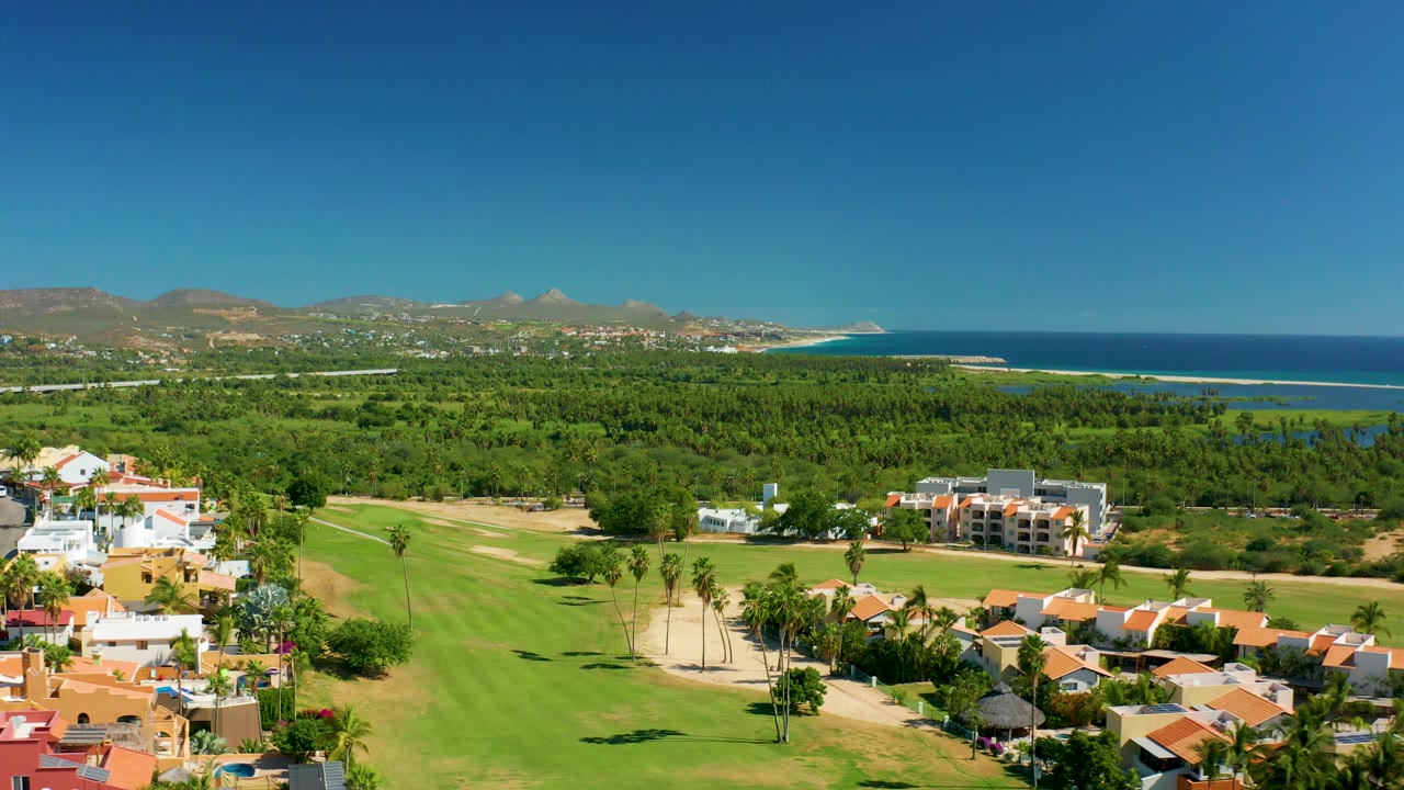Aerial panorama of green fairways, homes, palms, ocean, and mountains.
