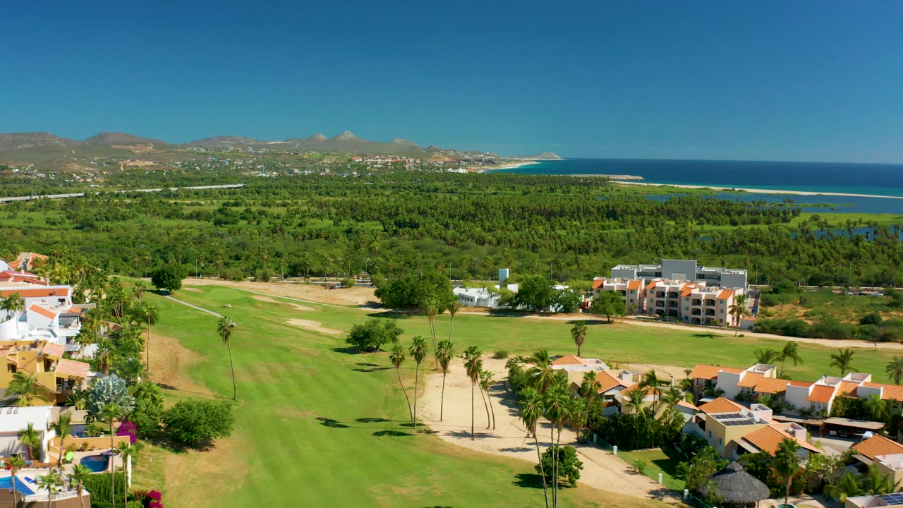 Opening aerial frame over San Jose del Cabo golf-course greens with palm trees and orange-roofed homes in the foreground, ocean and low mountains beyond, and no visible people.