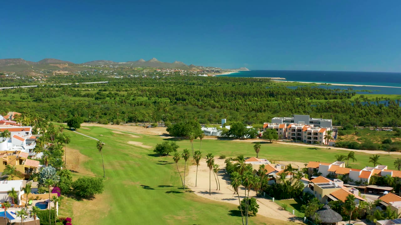 Second aerial frame shows the fairway and sand path below, a cluster of residential buildings to the right, dense coastal vegetation, ocean, mountains, and no visible people.