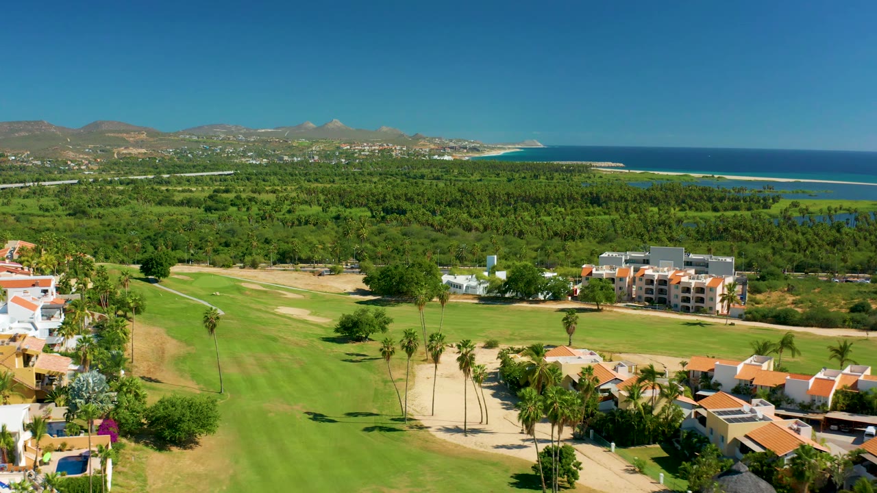 Third frame widens across San Jose del Cabo greenery, with manicured fairway below, apartment-style buildings at right, ocean and mountain horizon beyond, and no visible people.
