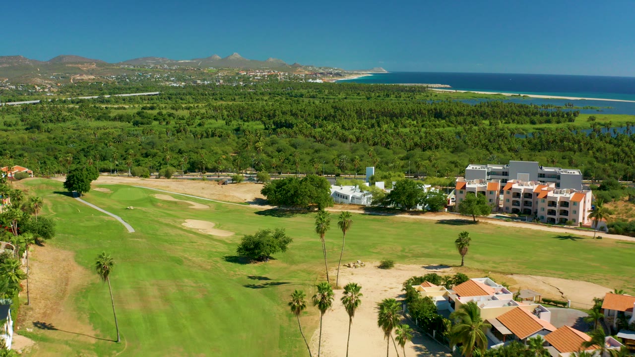 Opening aerial view over a San Jose Del Cabo golf course with green fairway, sand traps, palms, low coastal buildings, ocean, distant mountains, and no visible people.