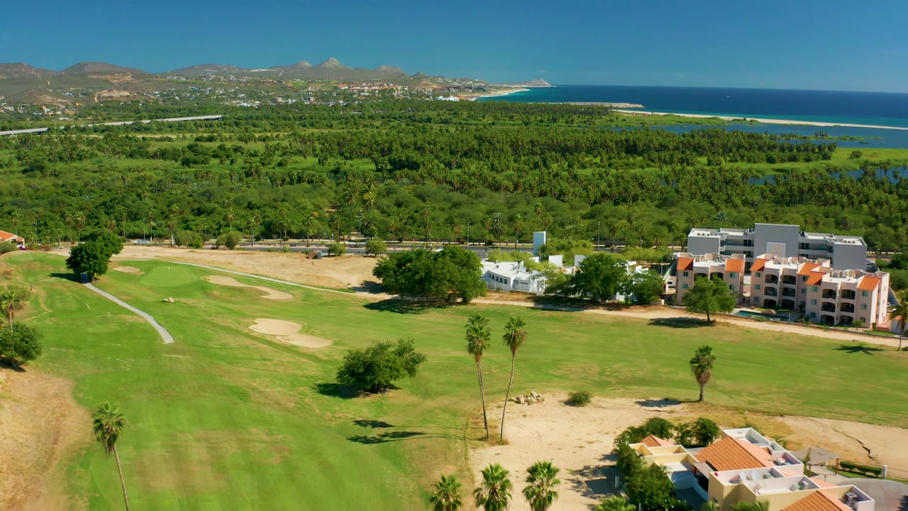 Aerial view across golf greens, sand traps, palms, buildings, ocean, and mountains.