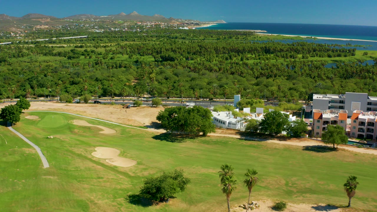 Opening aerial frame over a San Jose del Cabo golf course, with green fairway and sand traps in front, low buildings and trees beyond, ocean and hills in the distance, and no visible people.