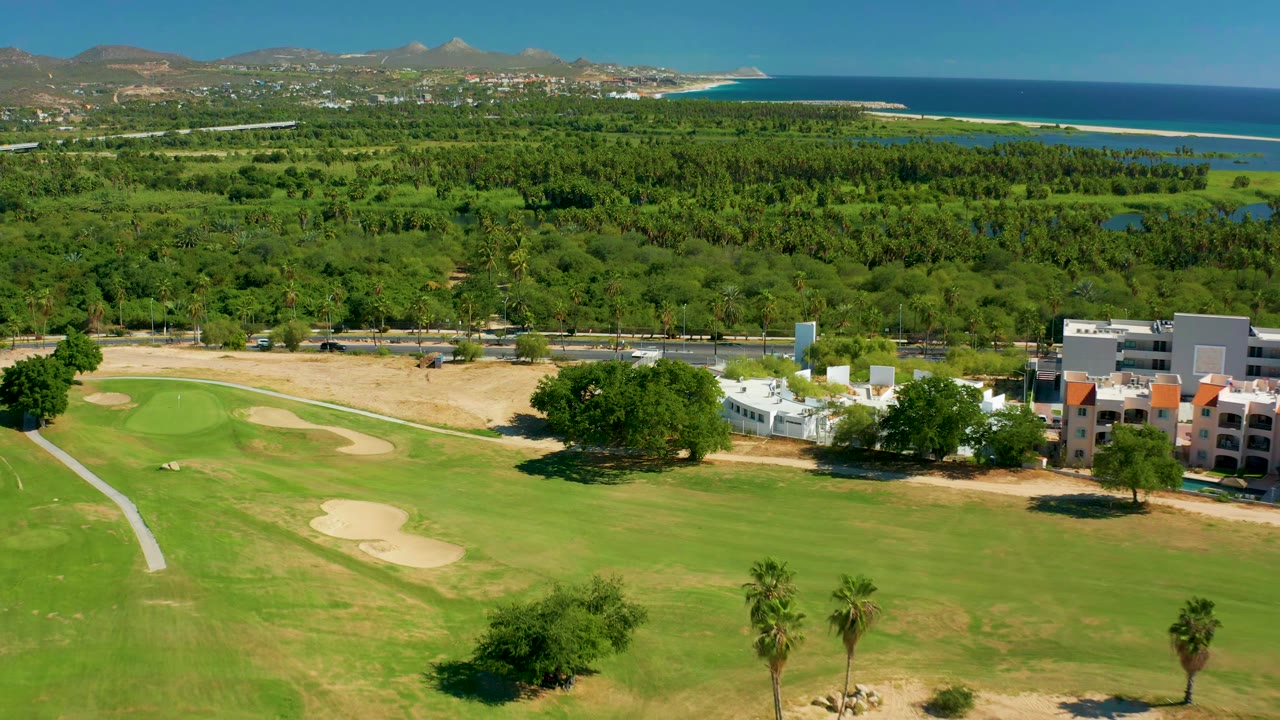 Aerial frame in San Jose del Cabo showing the fairway and bunkers slightly re-centered, with palms and low buildings near the course edge, coastline behind the tree canopy, and no visible people.