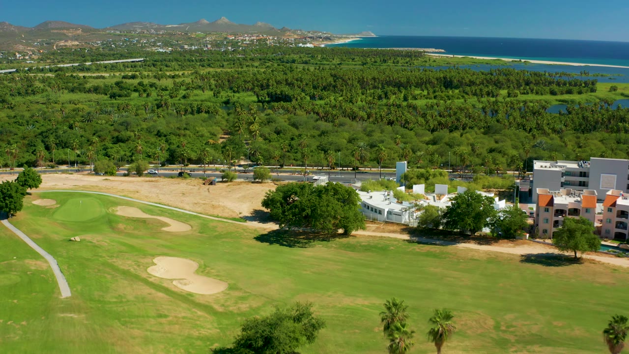 San Jose del Cabo aerial frame with the green and sand traps spread across the lower half, low buildings at right, a broad green landscape behind them, ocean at the horizon, and no visible people.