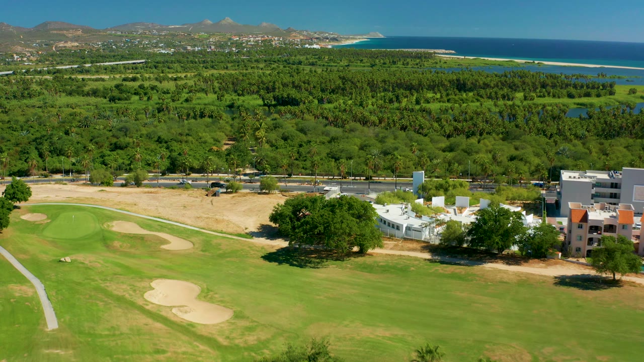 Aerial golf course scene with fairway, sand bunker, large tree, buildings, ocean, and hills.