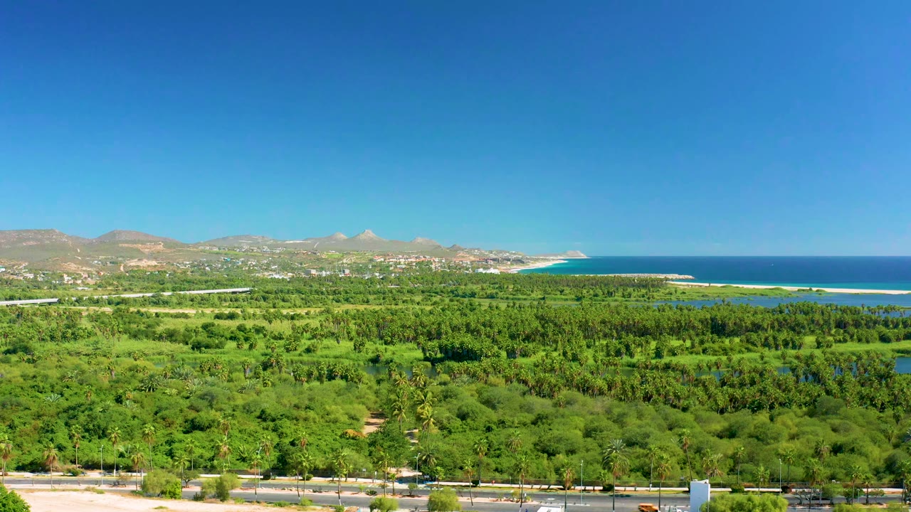 Early frame of San Jose Del Cabo showing dense green vegetation below, blue coastal water beyond, distant mountains, clear sky, and no visible people.