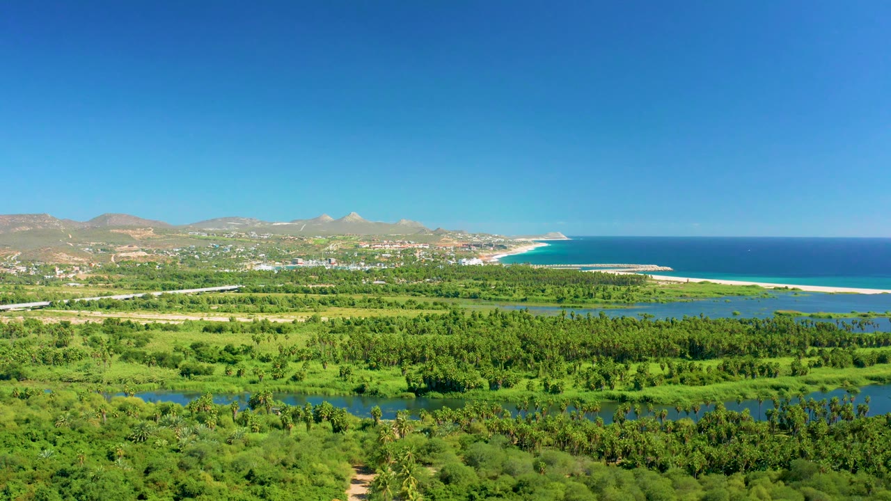 Fourth frame over San Jose Del Cabo places a pale road above the green valley, with river water, beach, ocean, dry mountains, open sky, and no visible people.